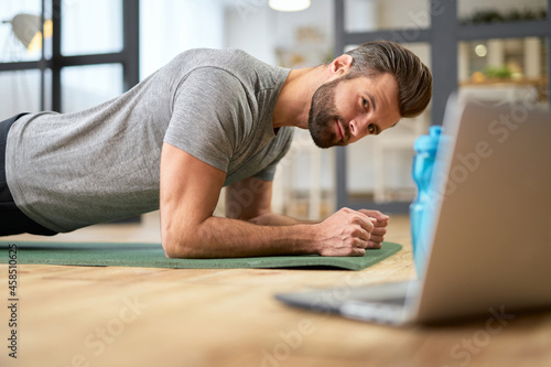 Bearded young man using notebook and doing plank at home