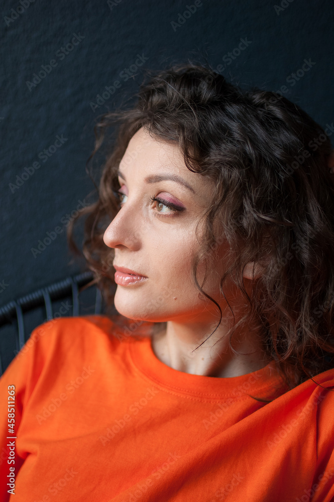 Fototapeta premium Female portrait of a beautiful curly girl in bright orange t-shirt at home in her apartment on dark background, happy people concept