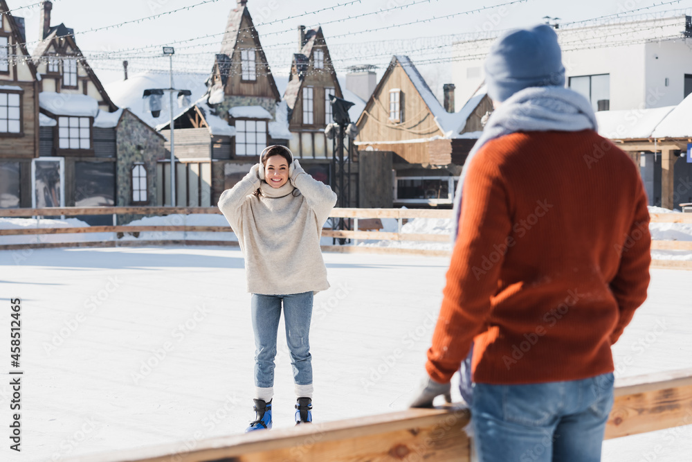 smiling young woman in ice skates looking at blurred boyfriend on foreground smiling young woman in ice skates looking at blurred boyfriend on foreground