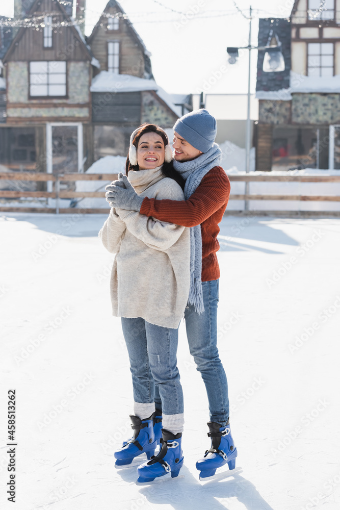 full length of happy man hugging young woman in ear muffs on ice rink full length of happy man hugging young woman in ear muffs on ice rink