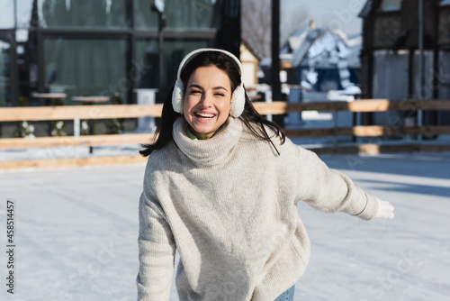 cheerful young woman in sweater and ear muffs skating on ice rink
