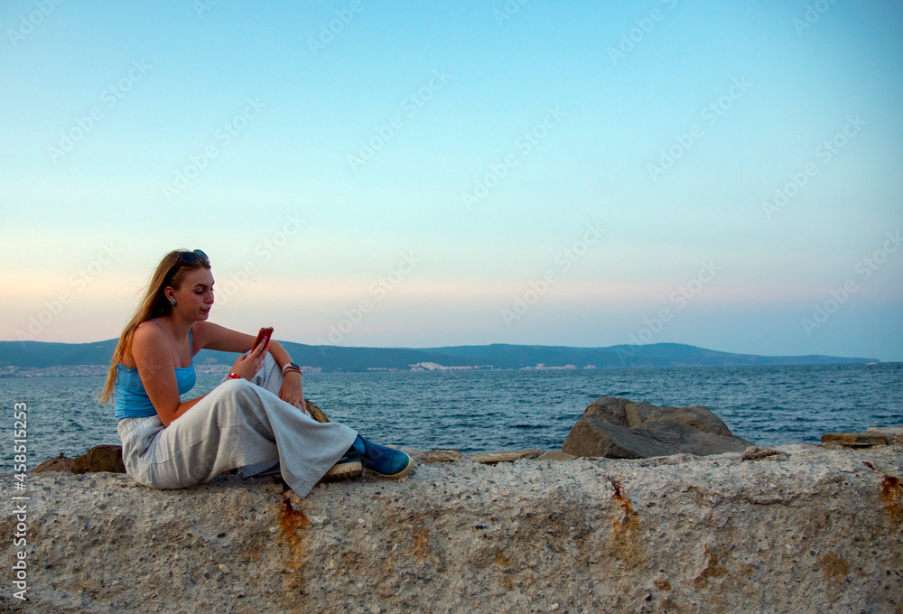 A blonde girl in dark glasses takes pictures of herself on her phone against the background of the sea .