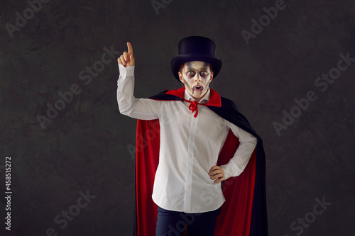 Child in vampire costume against dark studio background. Kid dressed as Dracula for Halloween party. Boy wearing top hat, red and black cape, white shirt, fake fangs and scary makeup points finger up