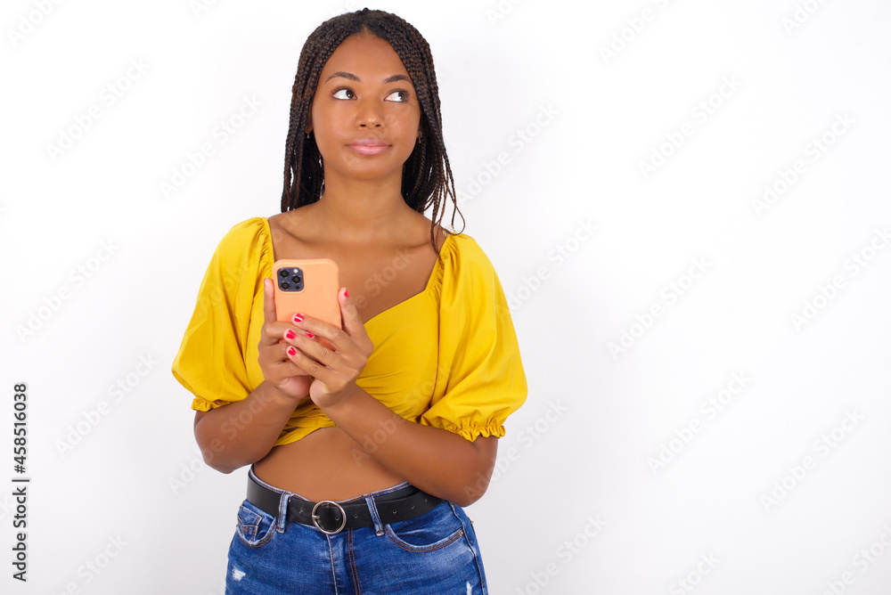 Portrait of afro american woman with braids wearing sexy yellow t-shirt on white wall with dreamy look, thinking while holding smartphone. Tries to write up a message. Portrait of afro american woman with braids wearing sexy yellow t-shirt on white wall with dreamy look, thinking while holding smartphone. Tries to write up a message.