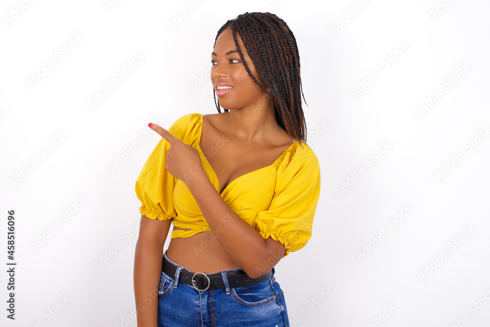 Happy cheerful smiling afro american woman with braids wearing sexy yellow t-shirt on white wall looking and pointing aside with hand. Copy space and advertisement concept. Happy cheerful smiling afro american woman with braids wearing sexy yellow t-shirt on white wall looking and pointing aside with hand. Copy space and advertisement concept.