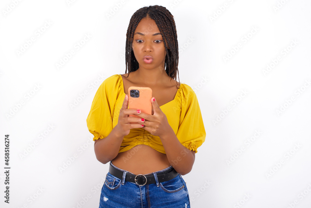 afro american woman with braids wearing sexy yellow t-shirt on white wall looks with bugged eyes, holds modern smart phone, receives unexpected message from friend, reads reminder. afro american woman with braids wearing sexy yellow t-shirt on white wall looks with bugged eyes, holds modern smart phone, receives unexpected message from friend, reads reminder.