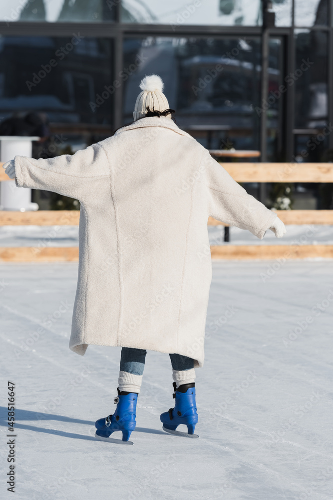 back view of young woman in winter hat and coat skating on ice rink