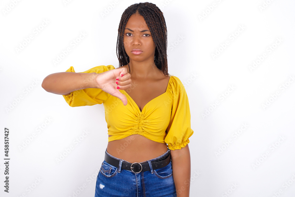 afro american woman with braids wearing sexy yellow t-shirt on white wall looking unhappy and angry showing rejection and negative with thumbs down gesture. Bad expression. afro american woman with braids wearing sexy yellow t-shirt on white wall looking unhappy and angry showing rejection and negative with thumbs down gesture. Bad expression.