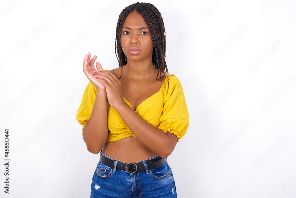 Surprised emotional afro american woman with braids wearing sexy yellow t-shirt on white wall rubs palms and stares at camera with disbelief Surprised emotional afro american woman with braids wearing sexy yellow t-shirt on white wall rubs palms and stares at camera with disbelief