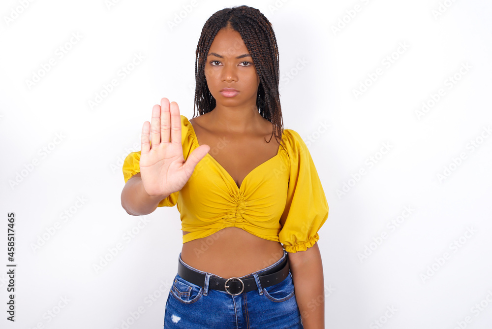 afro american woman with braids wearing sexy yellow t-shirt on white wall shows stop sign prohibition symbol keeps palm forward to camera with strict expression afro american woman with braids wearing sexy yellow t-shirt on white wall shows stop sign prohibition symbol keeps palm forward to camera with strict expression