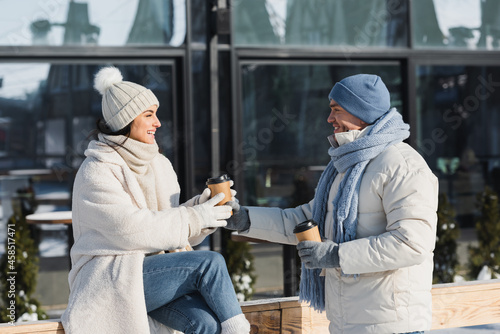 joyful young man giving paper cup to cheerful girlfriend in winter hat
