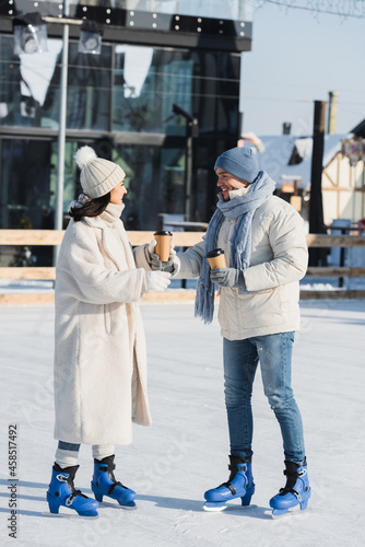 full length of happy young man giving paper cup to cheerful girlfriend in winter hat on ice rink