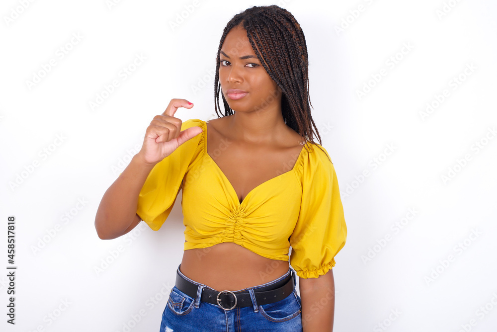 Displeased afro american woman with braids wearing sexy yellow t-shirt on white wall shapes little hand sign demonstrates something not very big. Body language concept. Displeased afro american woman with braids wearing sexy yellow t-shirt on white wall shapes little hand sign demonstrates something not very big. Body language concept.