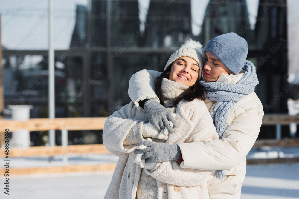 happy young man and woman in winter hats hugging on ice rink