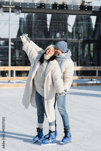full length of happy young man and woman in winter hats skating on ice rink