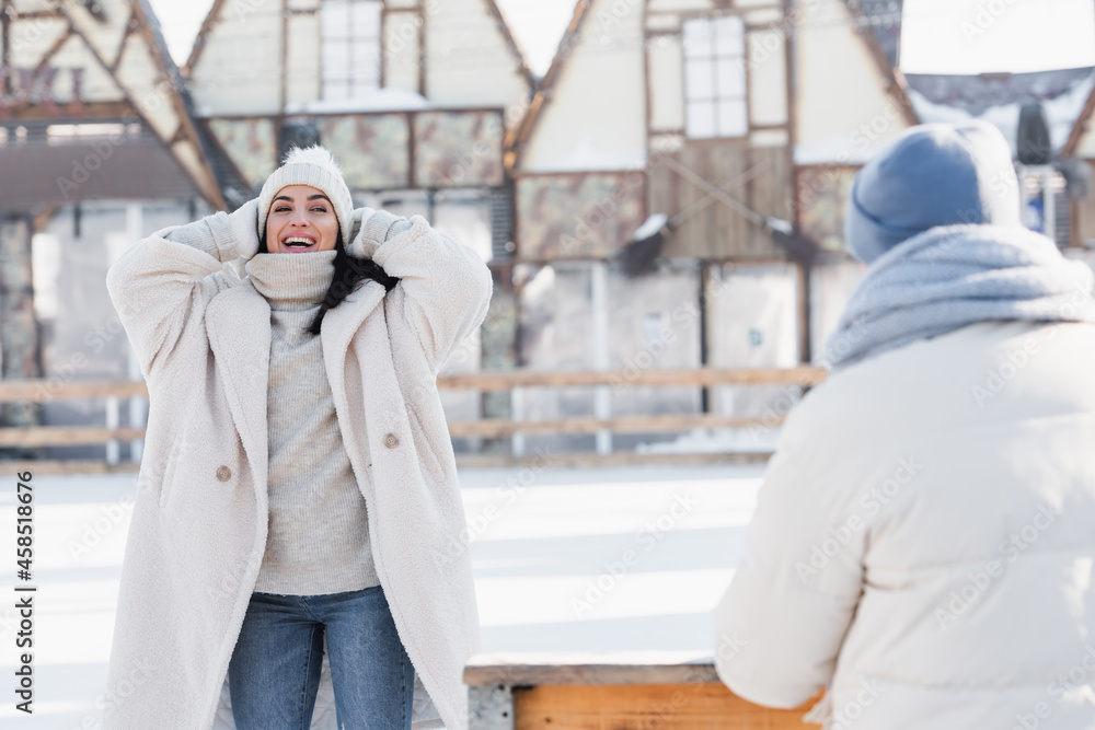 smiling young woman adjusting winter hat near blurred boyfriend