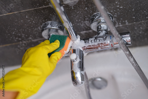 Housewife in rubber glove washing faucet in bathroom with sponge closeup