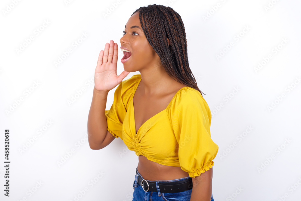 afro american woman with braids wearing sexy yellow t-shirt on white wall look empty space holding hand near her face and screaming or calling someone. afro american woman with braids wearing sexy yellow t-shirt on white wall look empty space holding hand near her face and screaming or calling someone.