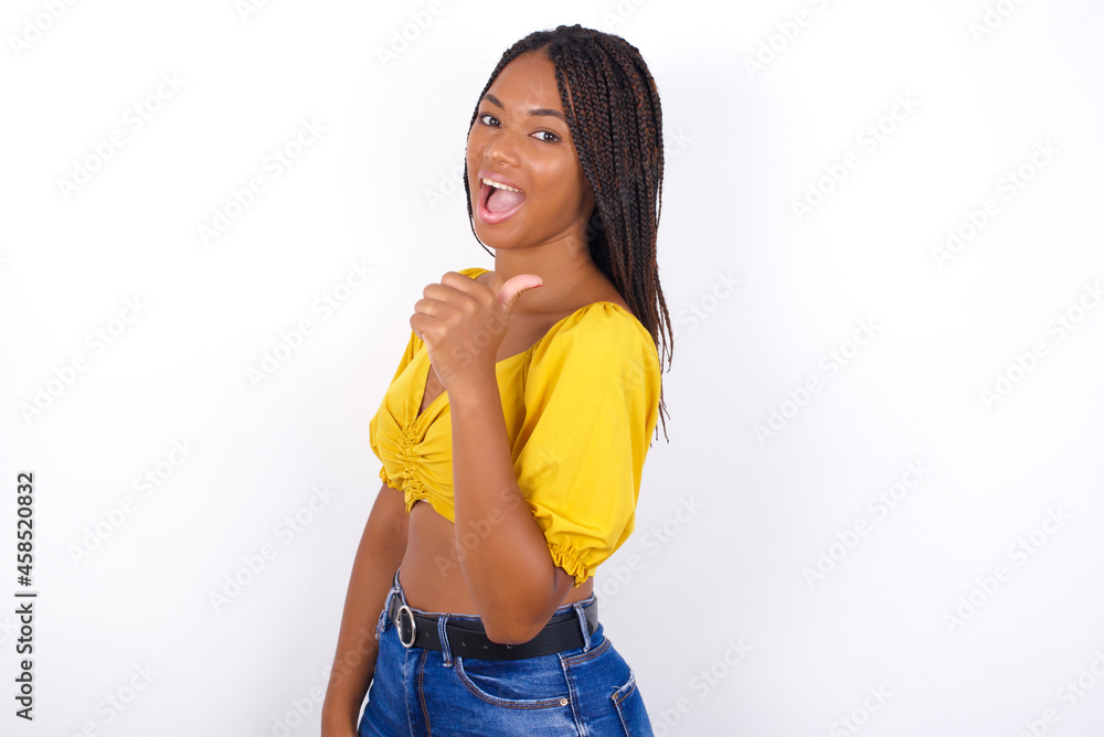 Impressed afro american woman with braids wearing sexy yellow t-shirt on white wall point back empty space Impressed afro american woman with braids wearing sexy yellow t-shirt on white wall point back empty space