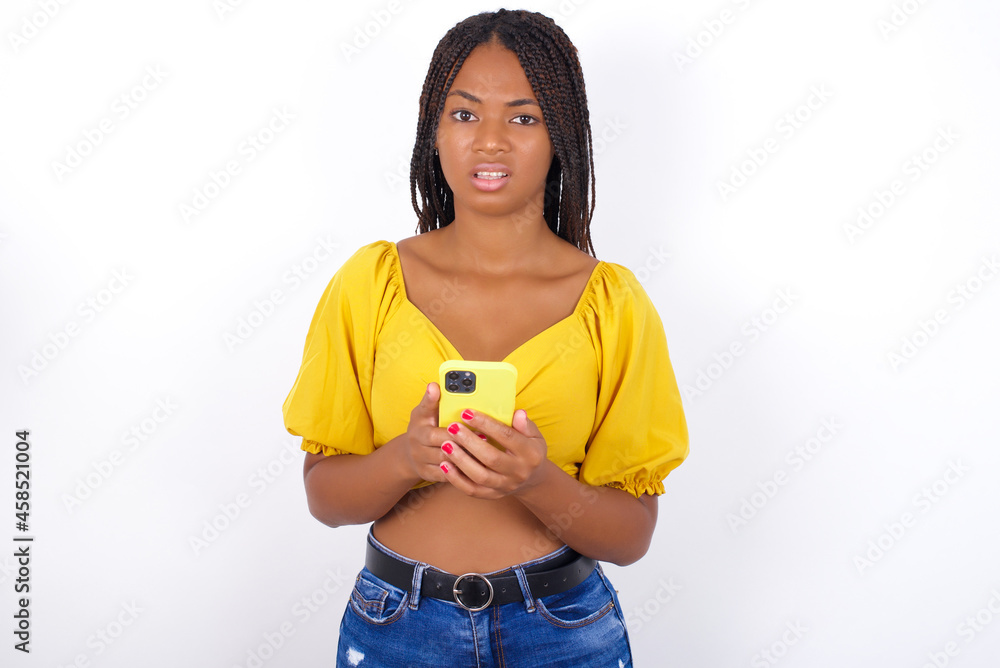 Photo of astonished crazy afro american woman with braids wearing sexy yellow t-shirt on white wall hold smartphone dislike feedback concept Photo of astonished crazy afro american woman with braids wearing sexy yellow t-shirt on white wall hold smartphone dislike feedback concept
