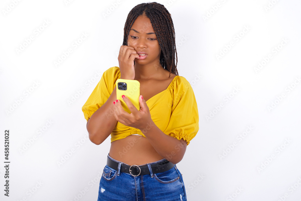 Portrait of pretty frightened afro american woman with braids wearing sexy yellow t-shirt on white wall chatting biting nails after reading some scary news on her smartphone. Portrait of pretty frightened afro american woman with braids wearing sexy yellow t-shirt on white wall chatting biting nails after reading some scary news on her smartphone.