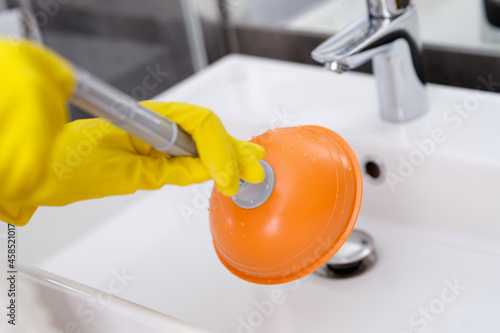 Plumber with rubber gloves cleaning sink with plunger in bathroom closeup