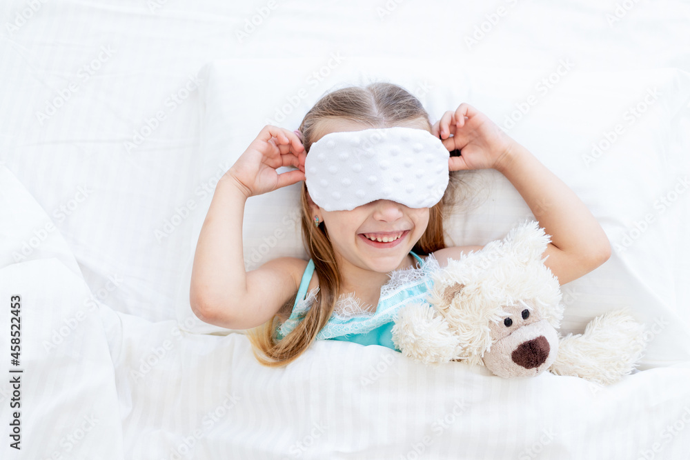 a little girl child with a sleep mask woke up in the morning at home on a bed on a white cotton bed with a teddy bear toy and a cute smile