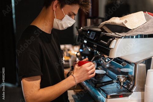 Man barista with a protective medical mask in a black T-shirt makes delicious coffee in a cafe.  epidemic and virus protection