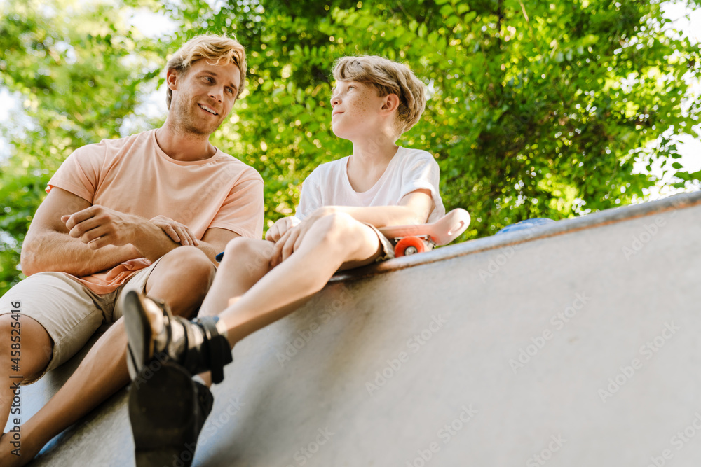 Ginger white man teaching his son how to ride skateboard on sports ground