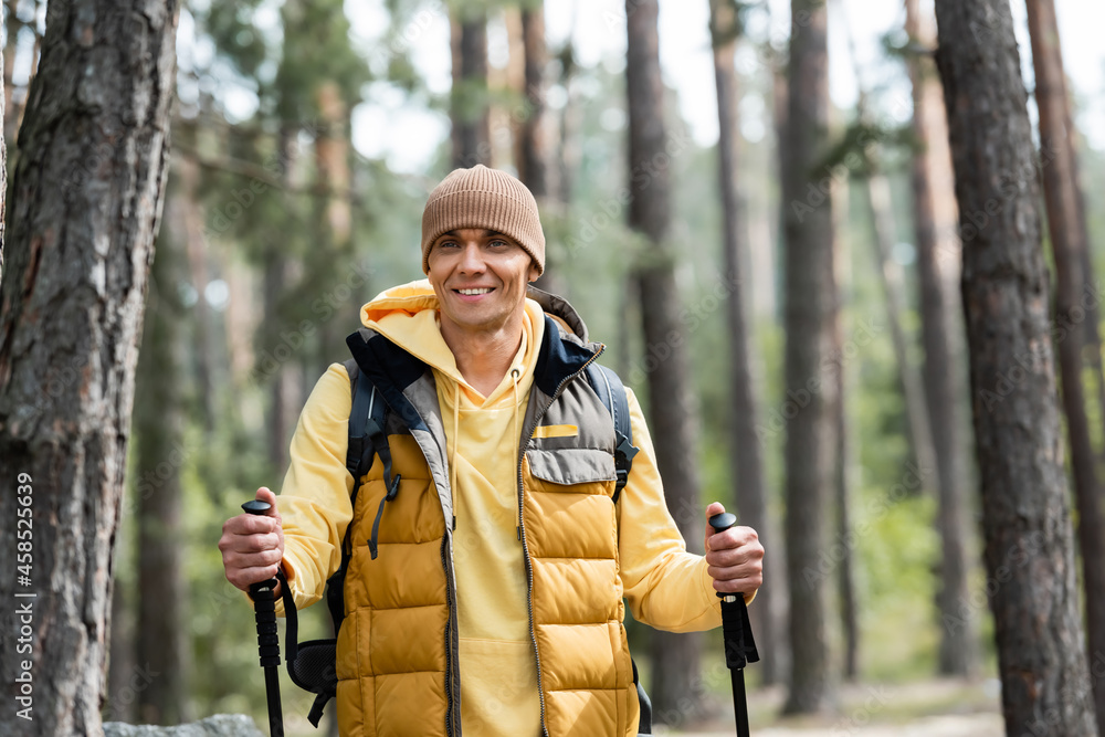 happy man in beanie and warm vest standing with trekking poles in forest happy man in beanie and warm vest standing with trekking poles in forest