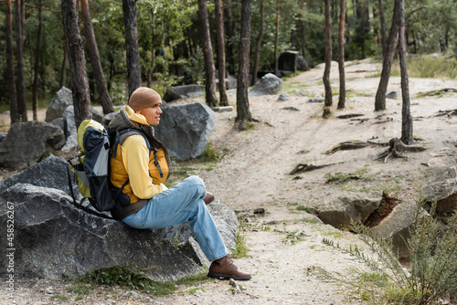 hiker with backpack sitting on stone in forest and looking away