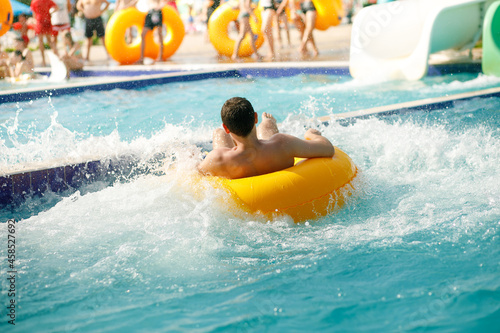 A cheerful man is having fun on a water slide in an amusement park with a yellow circle  glides with fun and water splashes around.