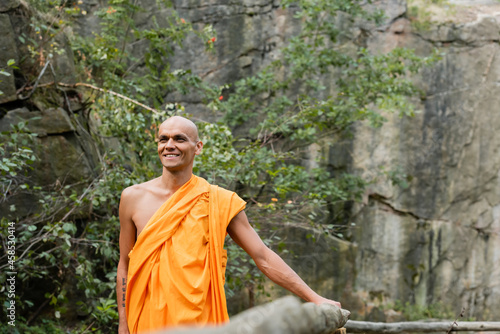 happy buddhist in orange kasaya walking near wooden fence in forest