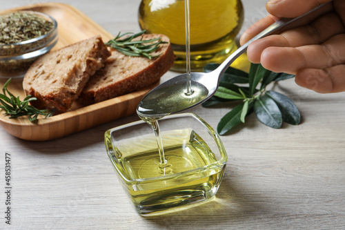 Woman pouring olive oil into spoon over bowl at wooden table, closeup