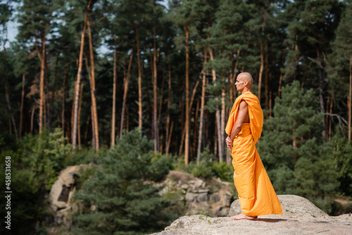 side view of barefoot buddhist monk in orange kasaya meditating on rocky cliff in forest photo