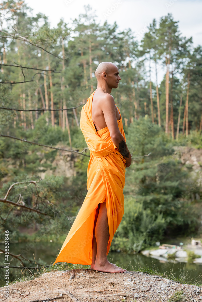 tattooed buddhist in traditional orange robe meditating on hill in forest tattooed buddhist in traditional orange robe meditating on hill in forest