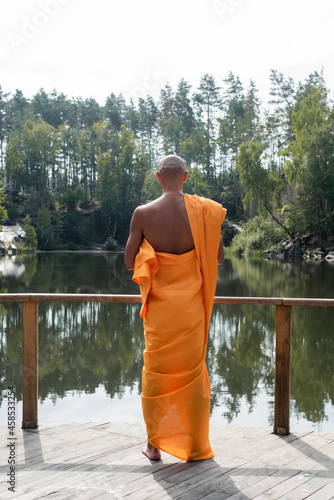 back view of buddhist in orange kasaya meditating near lake in forest photo