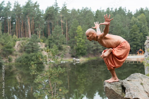 shirtless buddhist meditating in yoga pose with outstretched hands on rocky cliff over river