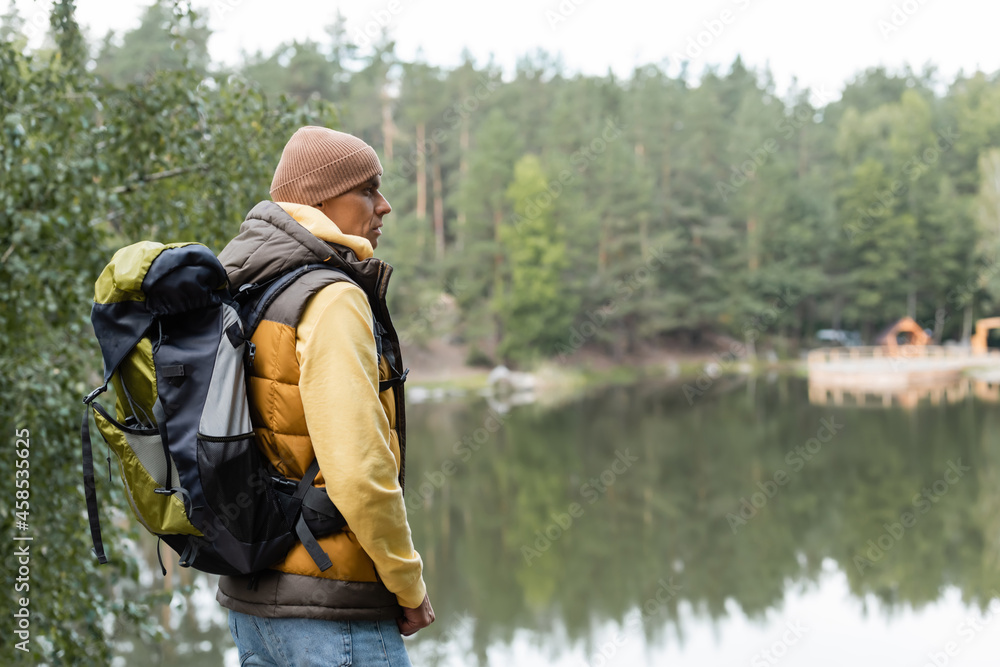 hiker with backpack looking away while standing near lake in forest hiker with backpack looking away while standing near lake in forest