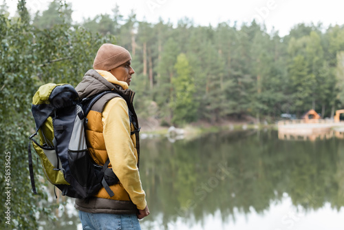 hiker with backpack looking away while standing near lake in forest
