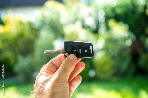POV male hand holding new car key against green defocused background with beautiful bokeh - three button device with open close and trunk signage