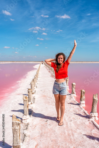 A beautiful young woman happily raises her hands up, on the shore of a salty pink lake with a blue sky. © KseniaJoyg