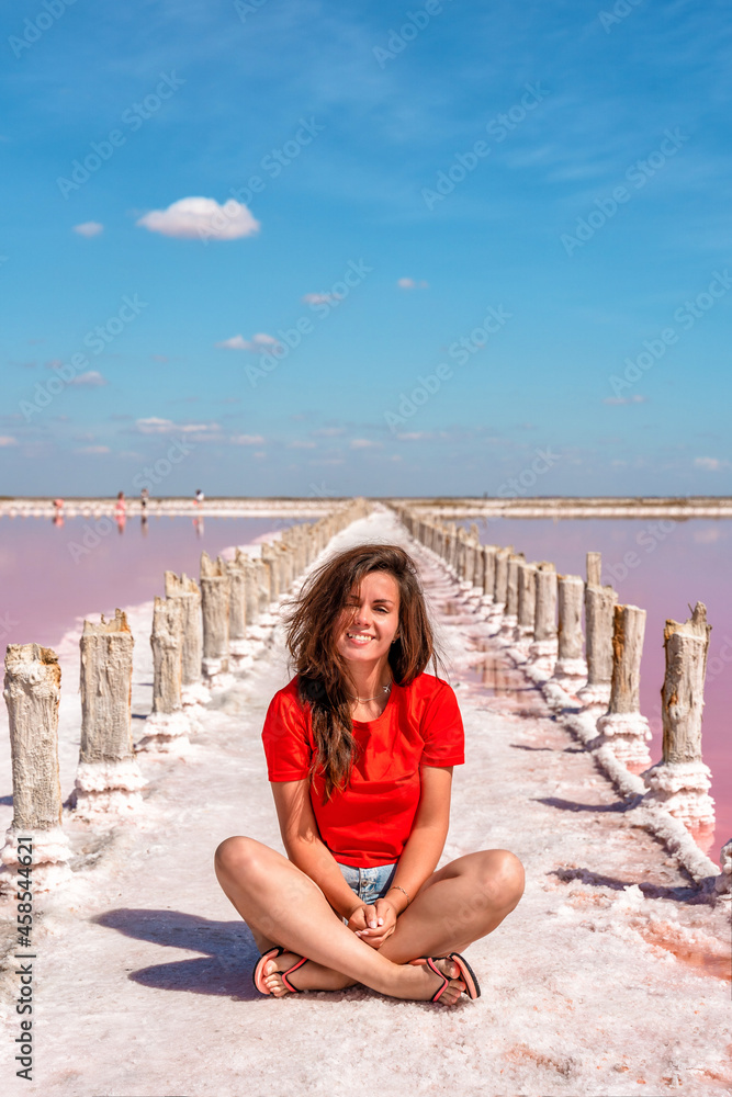 A cute young woman is sitting on a salty beach between wooden sticks on a salty pink lake with a blue sky. A peaceful landscape and relaxation