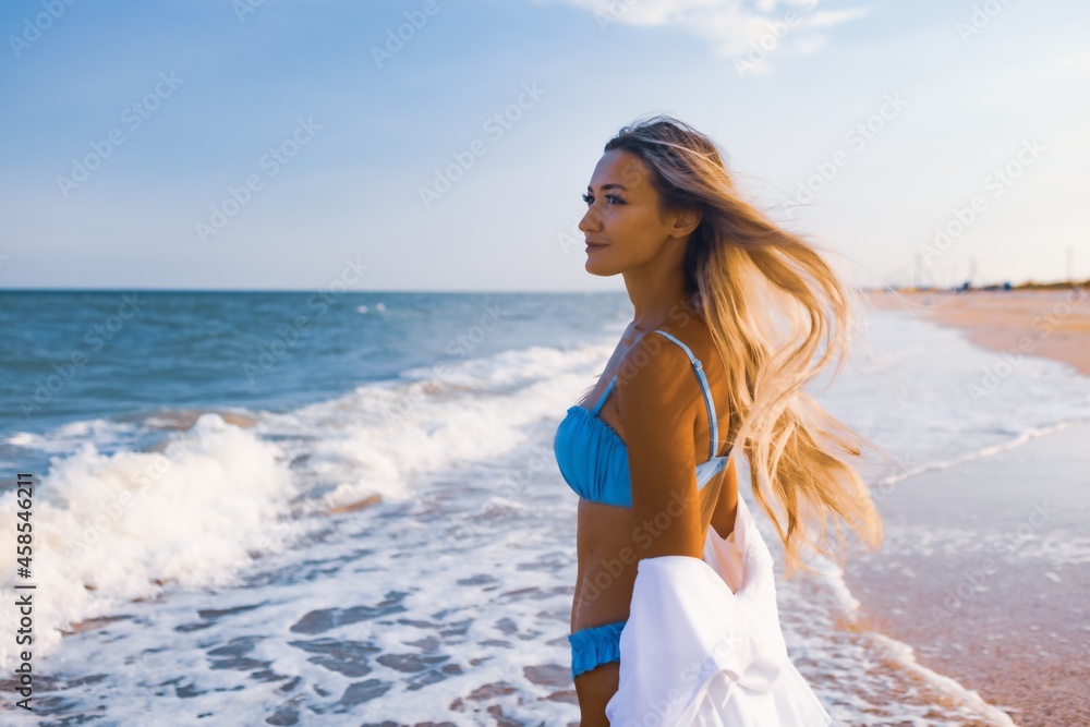 A slender girl in a gentle blue swimsuit and shirt walks along the sandy beach near the blue sea A slender girl in a gentle blue swimsuit and shirt walks along the sandy beach near the blue sea