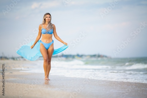 Obraz na płótnie A girl with blond hair in a bluish swimsuit and a bright shawl walks along the beach w salonie