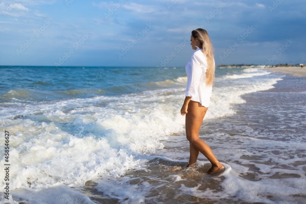 A slender girl in a gentle blue swimsuit and shirt walks along the sandy beach near the blue sea