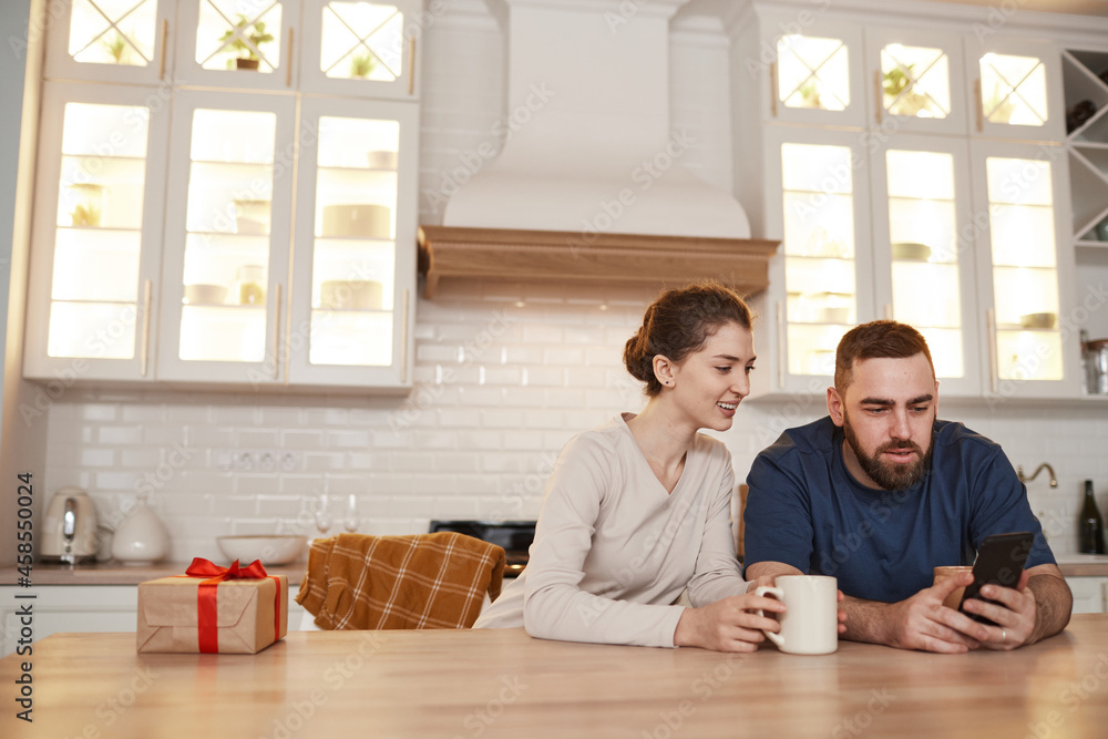 Cheerful young Caucasian couple sitting at table in kitchen and drinking coffee while surfing net together on smartphone Cheerful young Caucasian couple sitting at table in kitchen and drinking coffee while surfing net together on smartphone