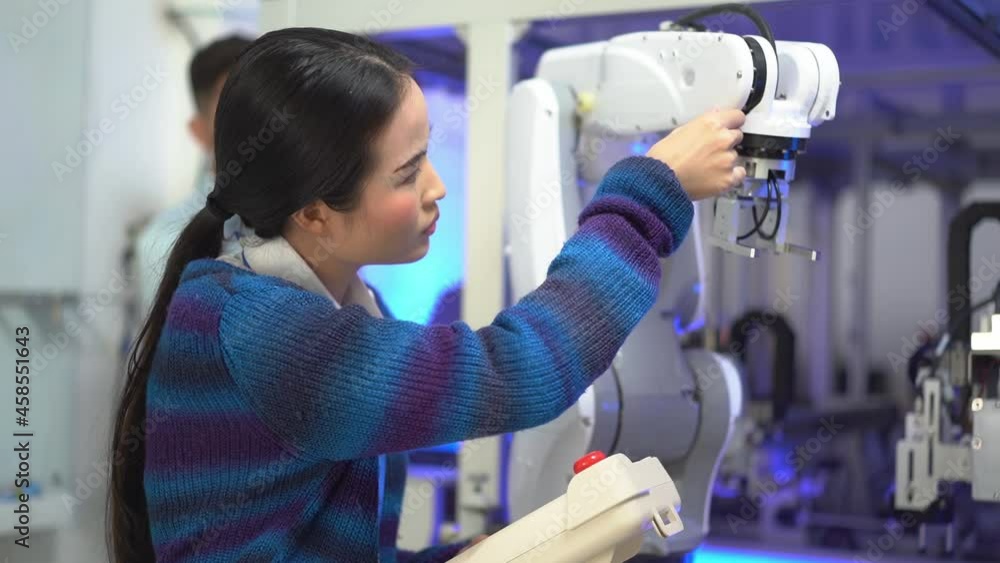 Asian female apprentice engineer in student uniform touching an automation robotic arm machine and testing the work of the machine with a robot controller while internship in an industrial factory. Asian female apprentice engineer in student uniform touching an automation robotic arm machine and testing the work of the machine with a robot controller while internship in an industrial factory.