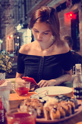 A beautiful girl reads messages on the phone while sitting in a cafe on the street in the evening. Lifestyle concept.