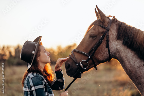 Beautiful smiling young woman in hat and shirt playing with brown horse in a field at sunset. Horseback riding. © sushytska